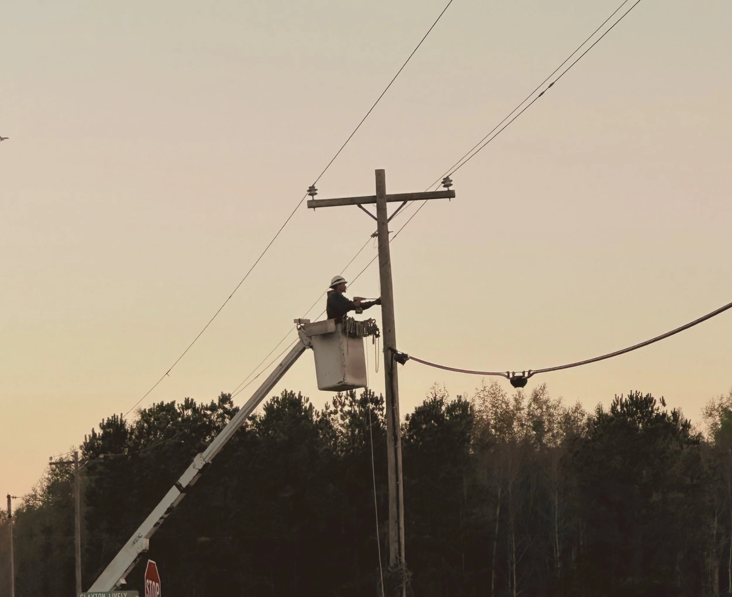 Worker in a bucket lift using a power drill on a utility pole against a sunset sky.