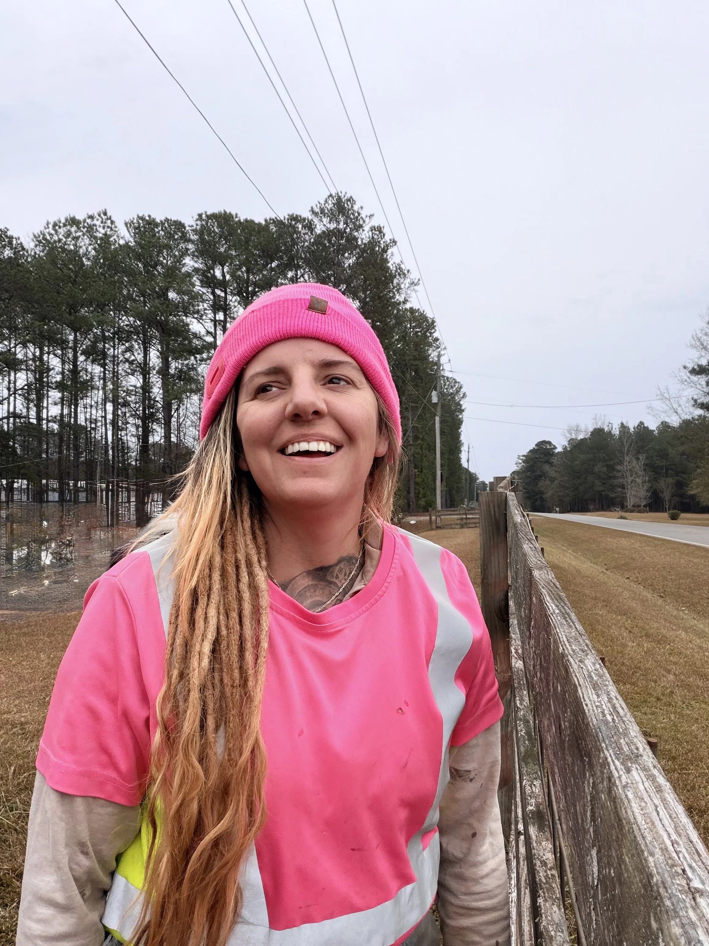A woman with long blonde hair, wearing a pink beanie and pink reflective safety vest, smiles outdoors next to a wooden fence, with trees and an overcast sky in the background.