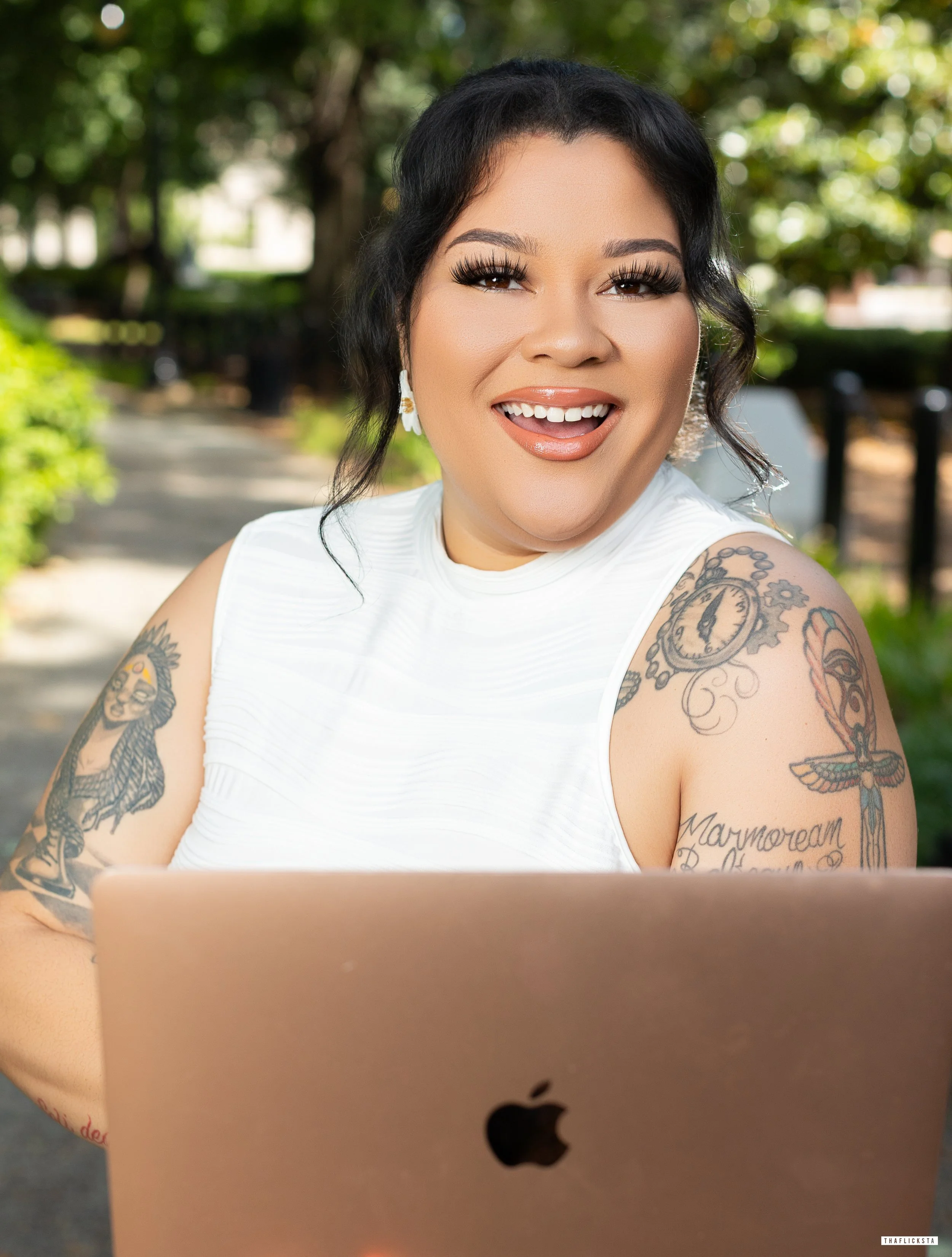 A woman with dark hair, tattoos on arms, wearing a white sleeveless top, smiling while looking at a laptop outdoors in a park.