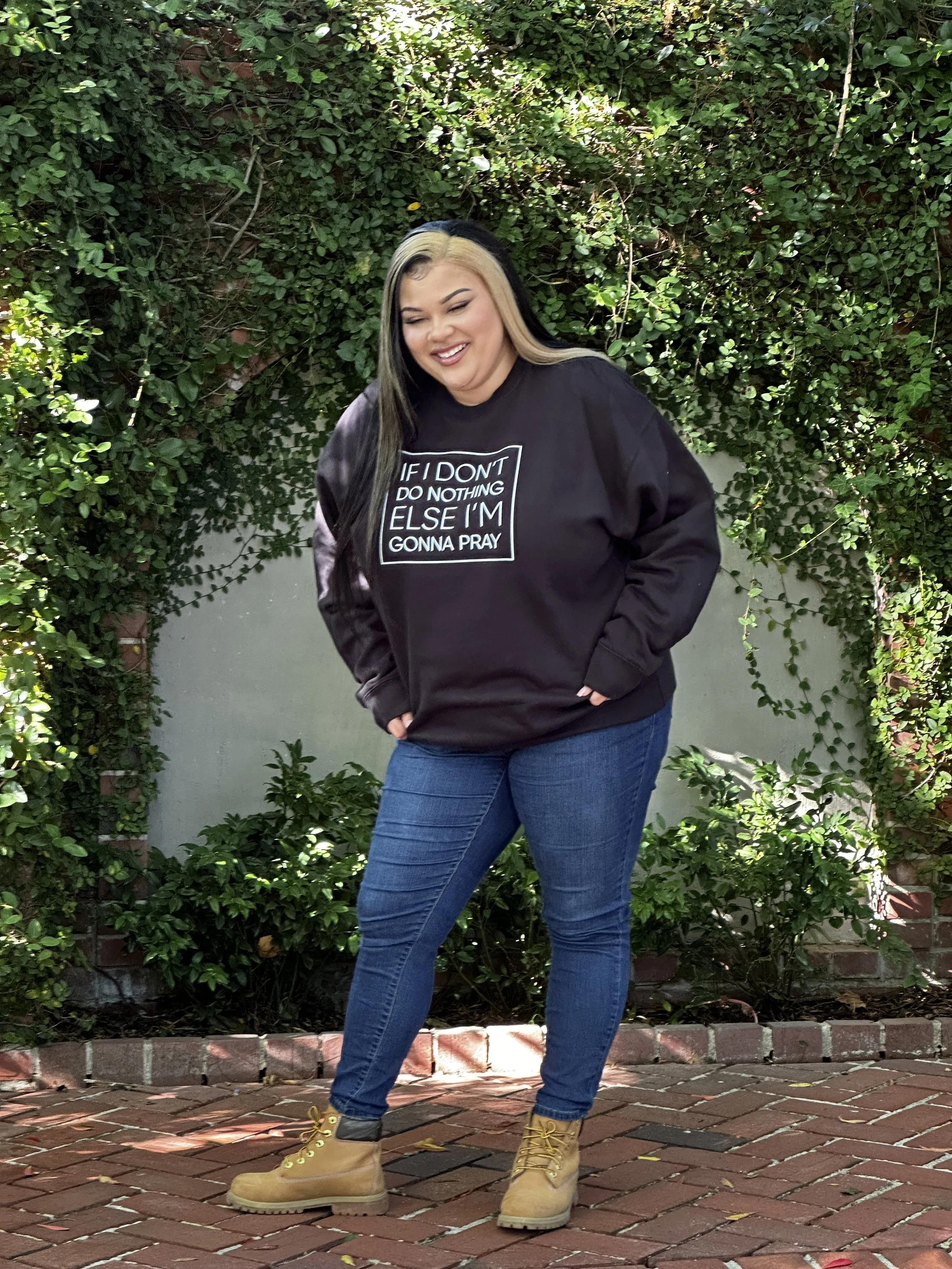 A woman with blonde and black hair, smiling, wearing a black sweatshirt with white text, blue jeans, and tan boots, standing outdoors on a brick pathway with greenery and ivy-covered wall in the background.