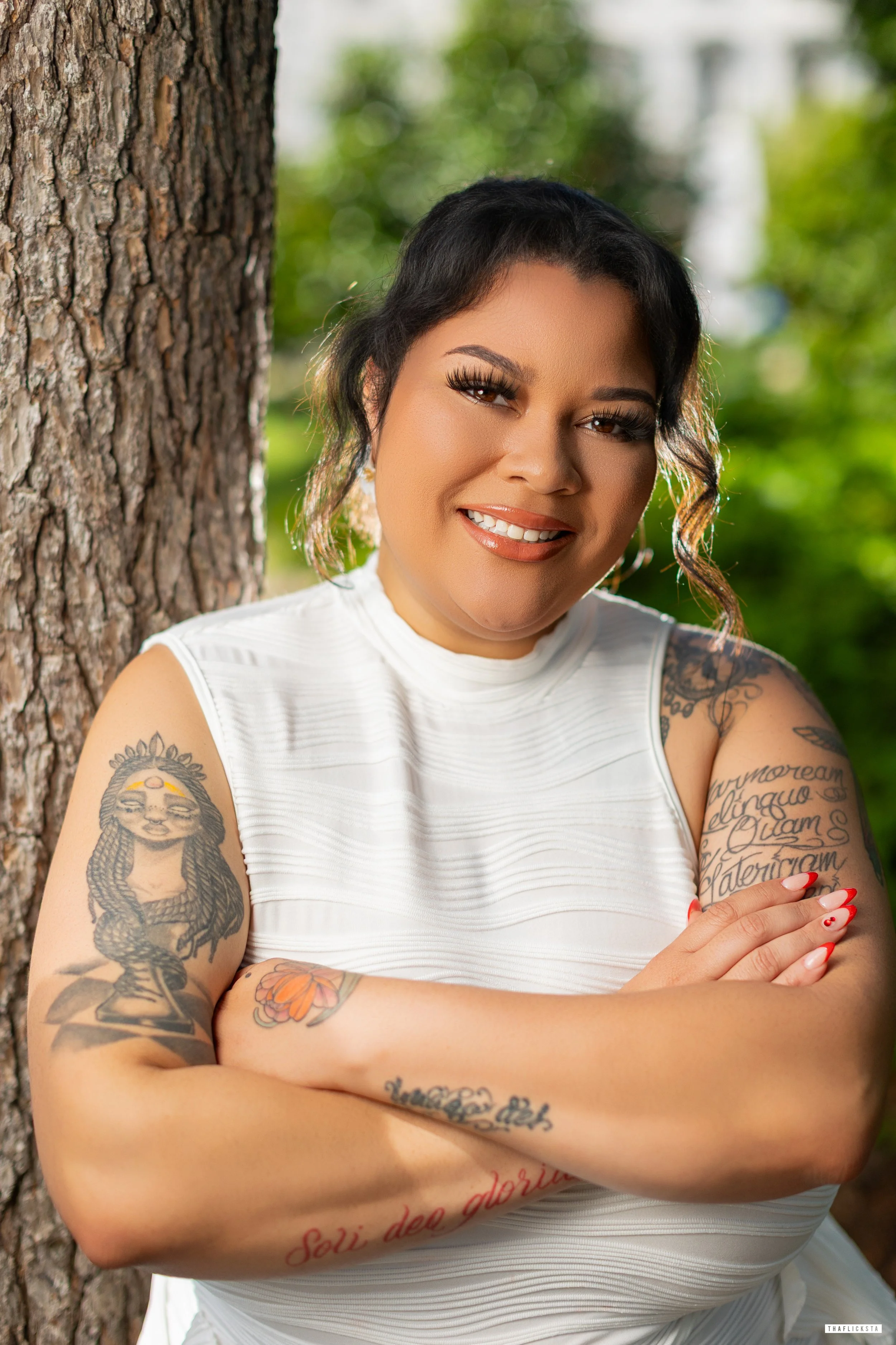 A woman with dark hair and tattoos on her arms, standing outdoors next to a tree with a green blurred background, smiling and crossing her arms.