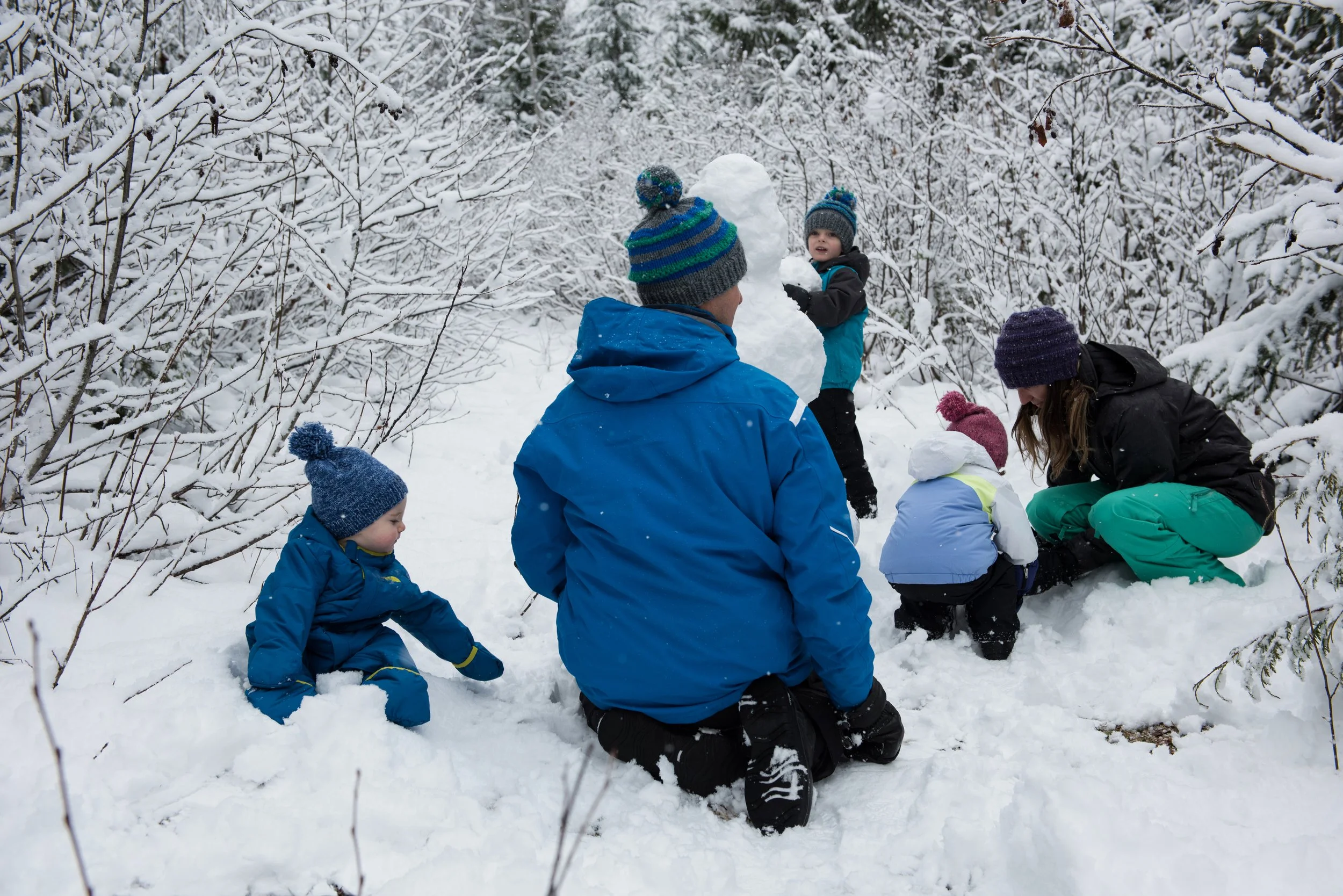 Family building a snowman and playing in snow in a wooded area.