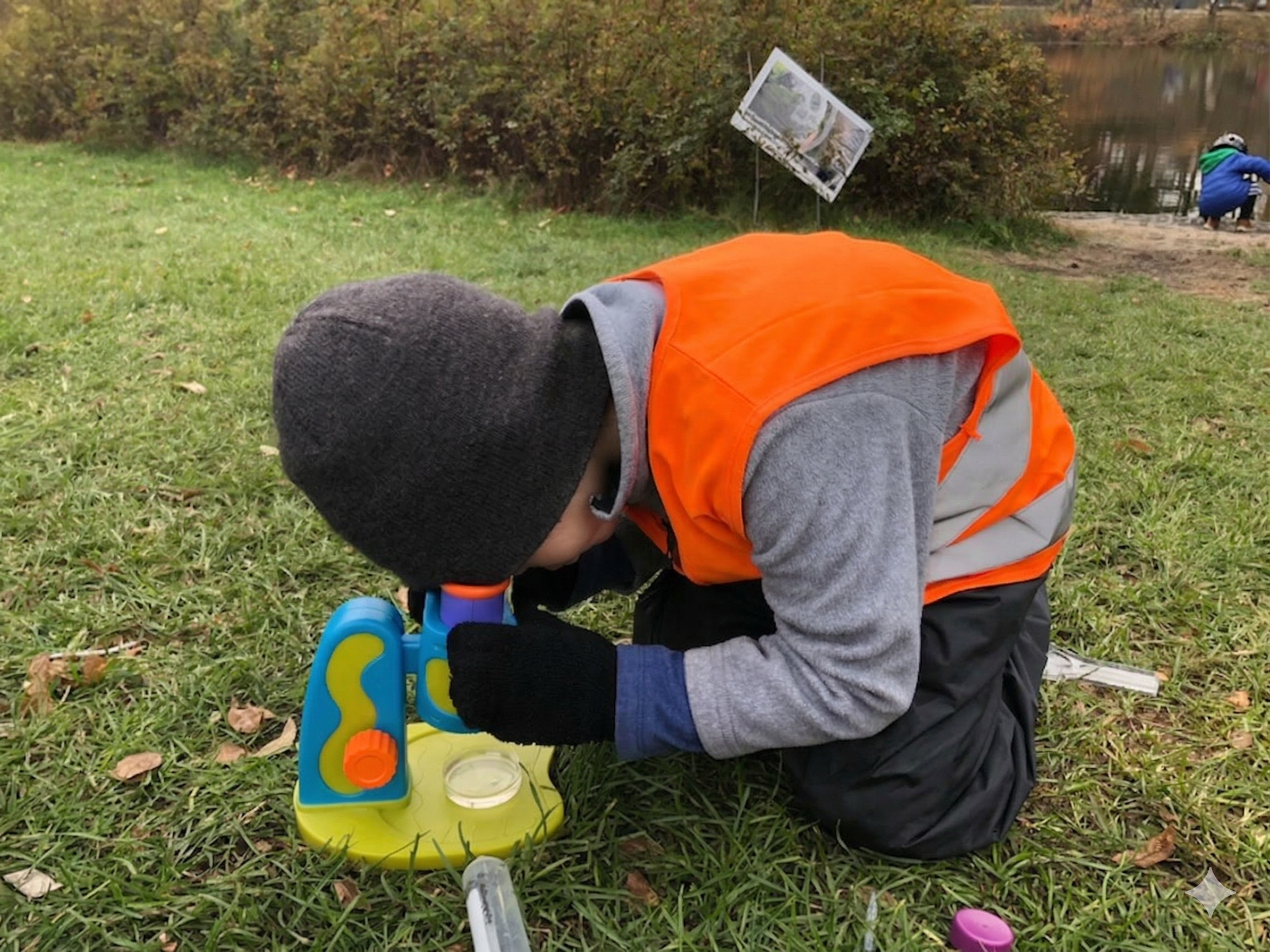 A young boy kneeling on green grass by a body of water, peering into a colorful microscope.  He is attending nature preschool.