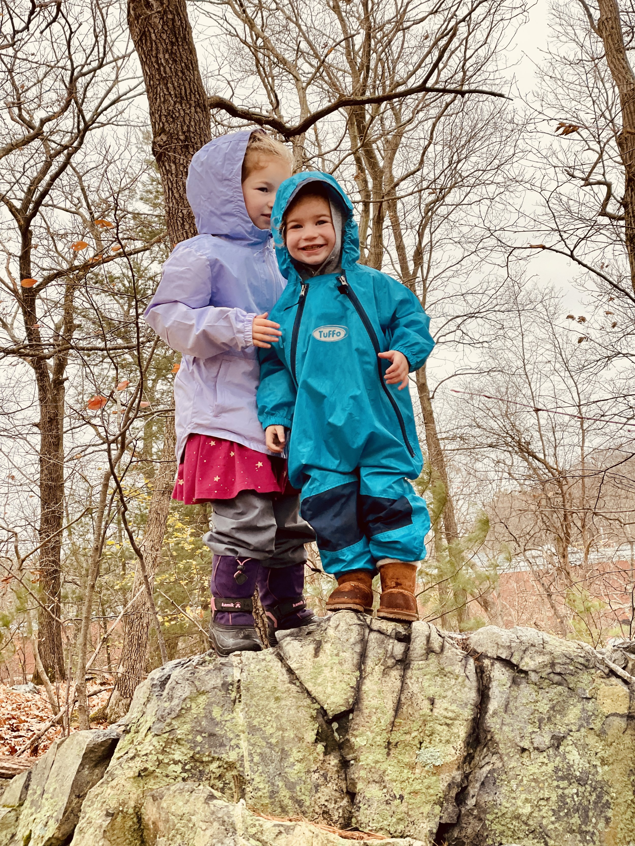 Two children standing on a large rock in a wooded area during fall, dressed in colorful rain jackets and boots, smiling and looking at the camera.
