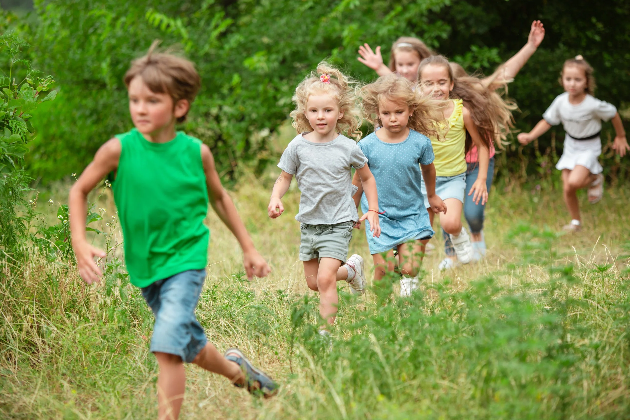 Seven children running through a grassy, wooded area outside