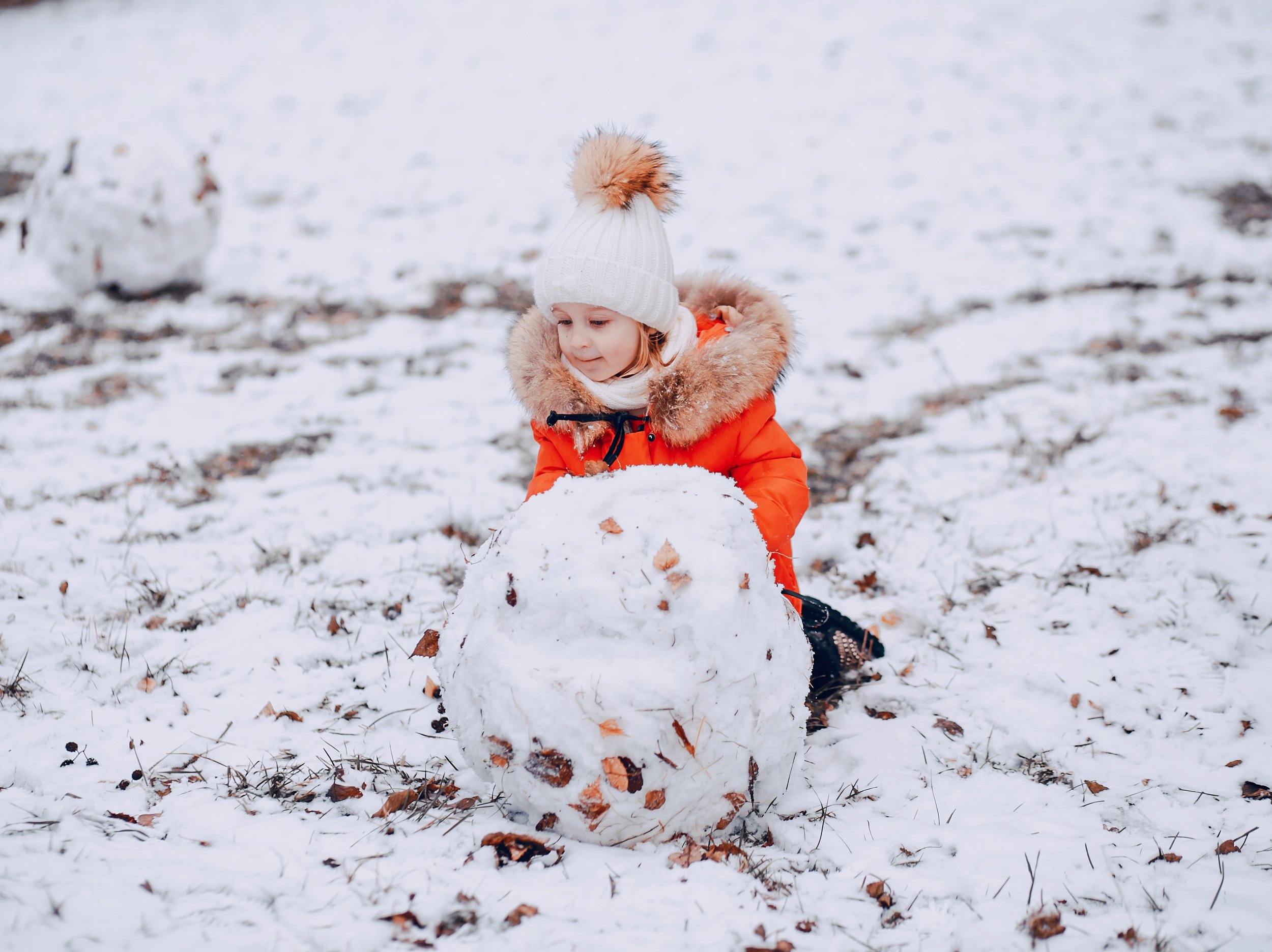 A young child in a white knit hat with a pom-pom, red winter coat with fur trim, is playing outside in the snow and building a snowman.