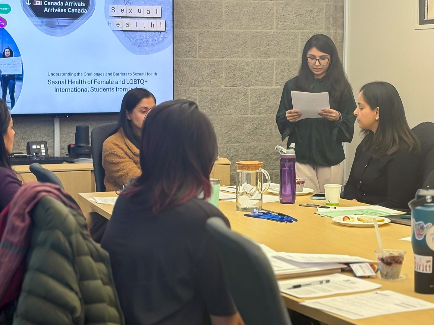 A woman is standing and speaking in a meeting room with others seated around a table. The room has a large screen displaying a presentation on sexual health of female and LGBTQ+ international students. The table has drinks, papers, and pens.