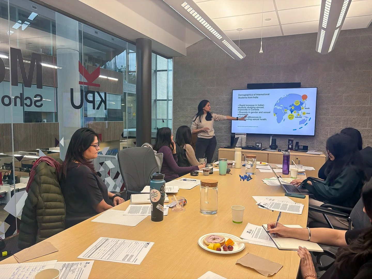 A woman presents a slideshow to a group of women seated around a conference table in a modern office conference room.