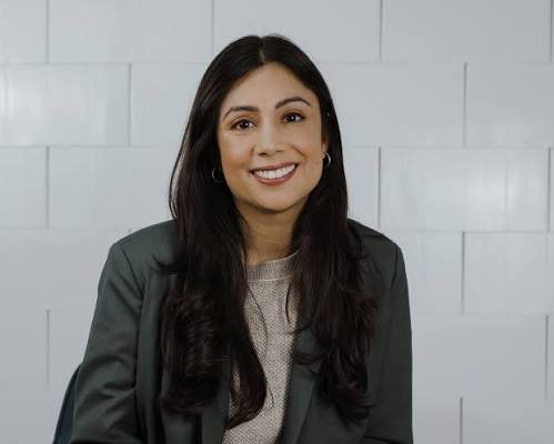 A woman with long dark hair, wearing a gray blazer and beige top, smiling and seated against a light-colored tiled wall.