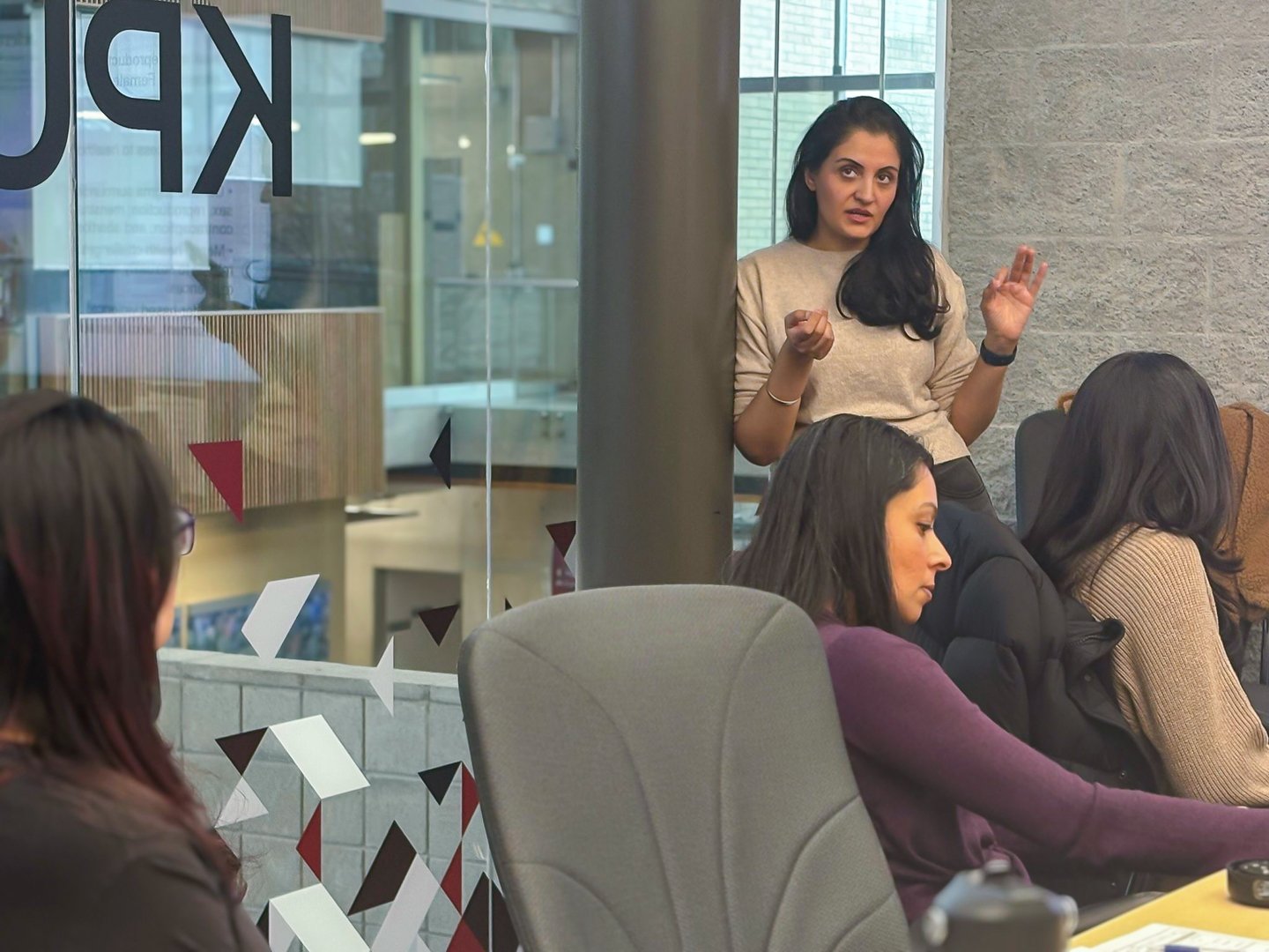 A woman standing and speaking to a group of women seated at a conference table in a modern office setting.