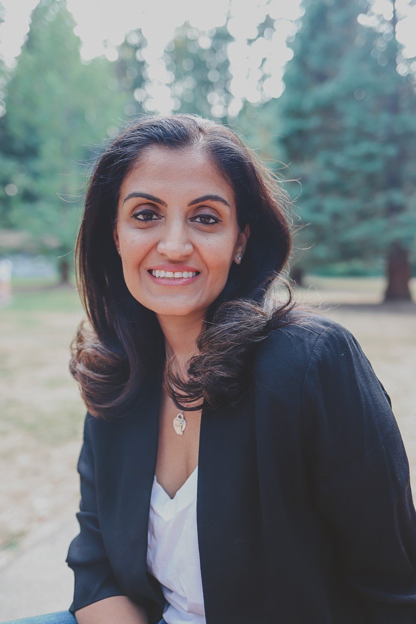 A South Asian woman with dark brown, shoulder-length hair, smiling outdoors in a park with greenery and trees in the background, wearing a black blazer, white top, and a small necklace.