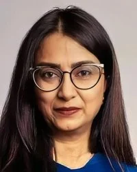 A South Asian woman with long dark hair wearing glasses and a black blazer, smiling slightly at the camera.