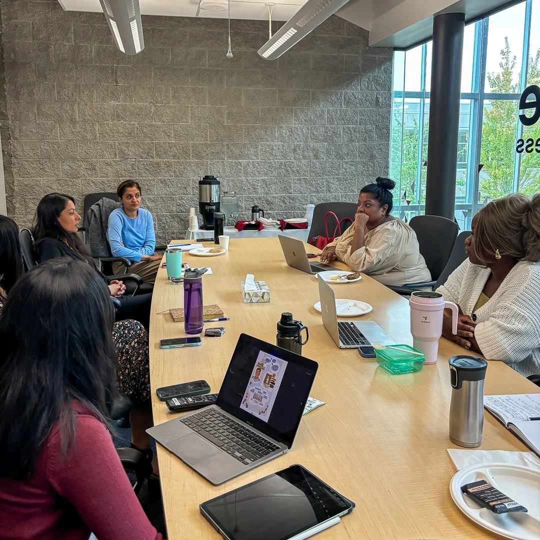 A diverse group of women seated around a conference table engaged in a meeting, with laptops, tablets, and coffee mugs on the table, inside a modern office with large windows and a brick wall.