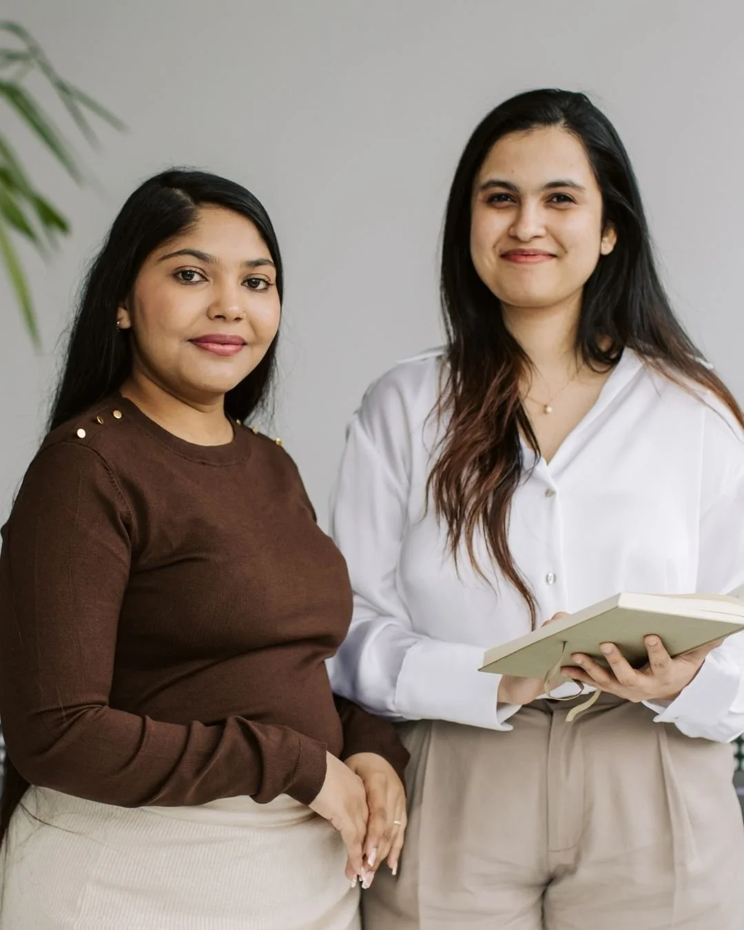 Two South Asian women standing together, one holding a notebook, smiling at the camera against a plain background.