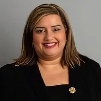 A woman with light brown hair, smiling, wearing a black blazer and a flower-shaped pin.