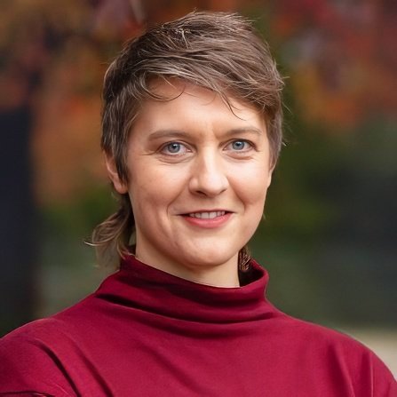 A woman with short brown hair and blue eyes, smiling, wearing a red turtleneck sweater outdoors with fall foliage in the background.