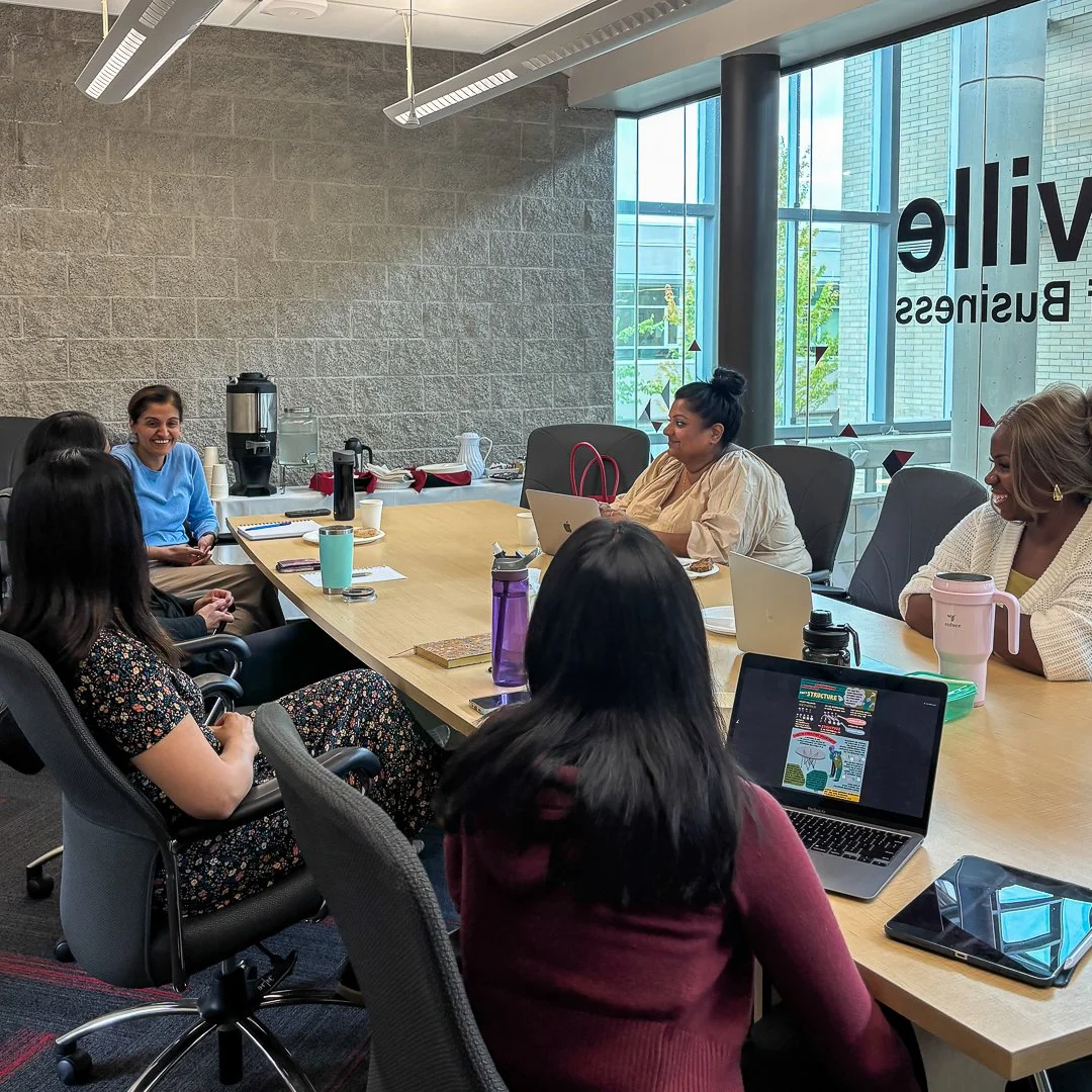 A diverse group of six women sitting around a conference table in a modern office, engaging in a discussion or meeting, with laptops, notebooks, and beverages on the table, large windows in the background letting in natural light.