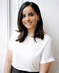 A South Asian woman with shoulder-length dark hair and a white top, smiling, standing indoors.