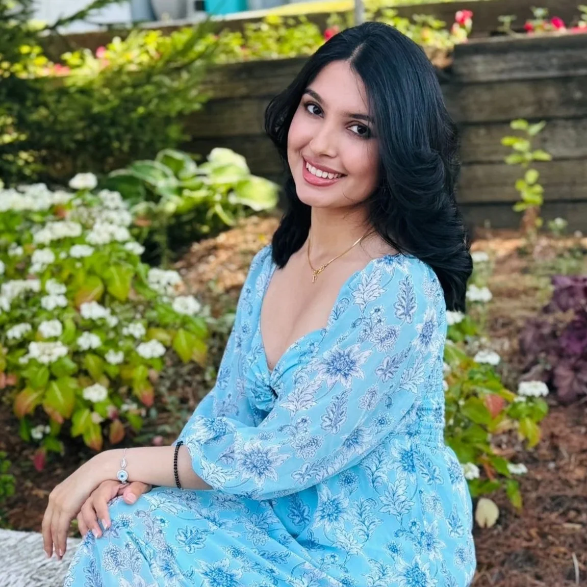 A young woman with long black wavy hair smiling, wearing a light blue floral dress, sitting outdoors in a garden with green plants and white flowers.