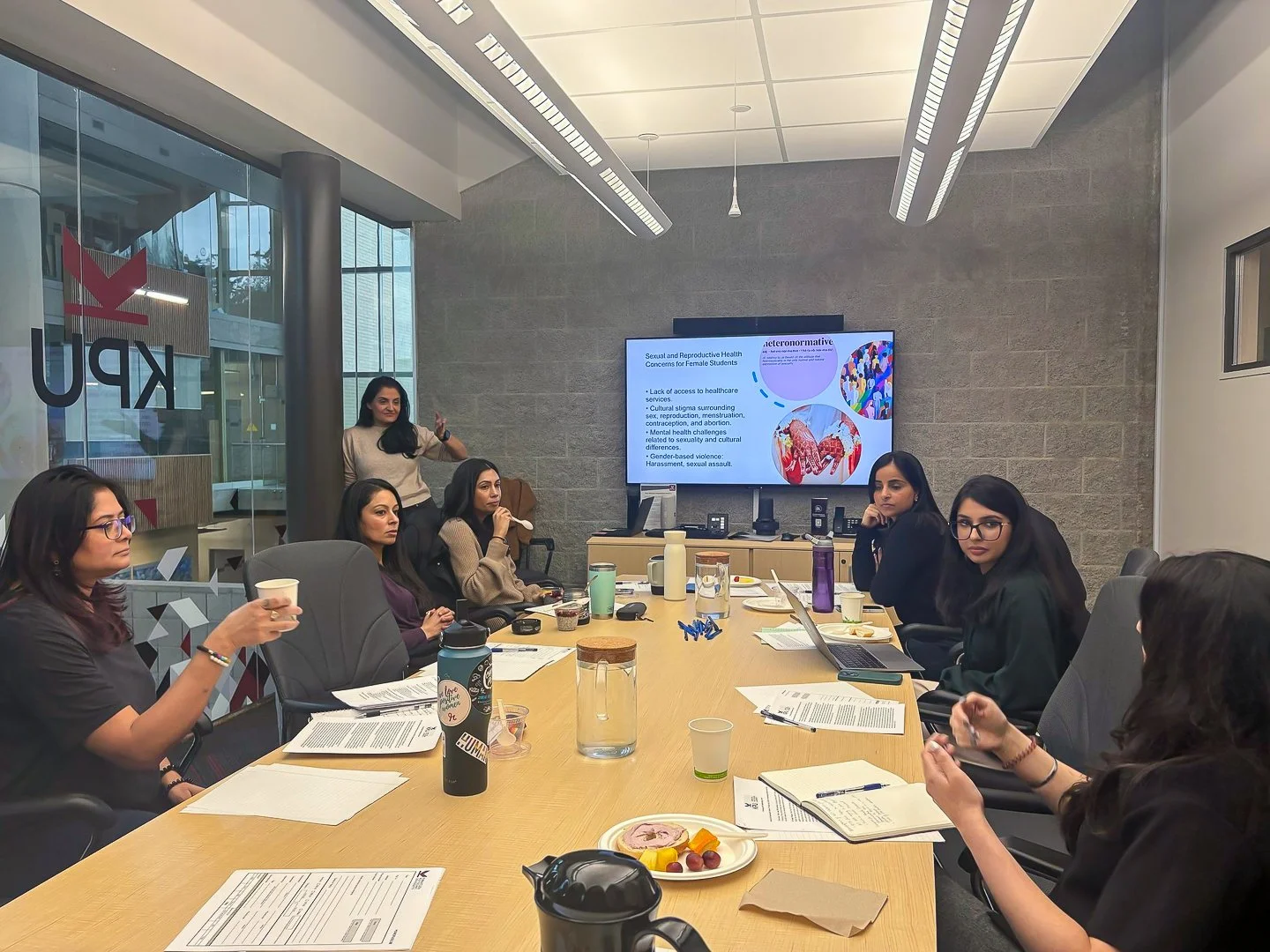 A group of women attending a presentation in a conference room, with some taking notes and others listening, while a woman stands near a large screen displaying a slide about sexual and reproductive health.