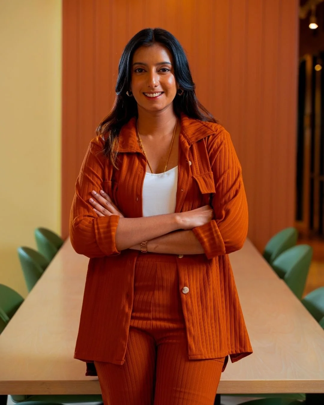 Young South Asian woman with black hair smiling, wearing an orange blazer and pants with a white top, standing with arms crossed in a modern office space