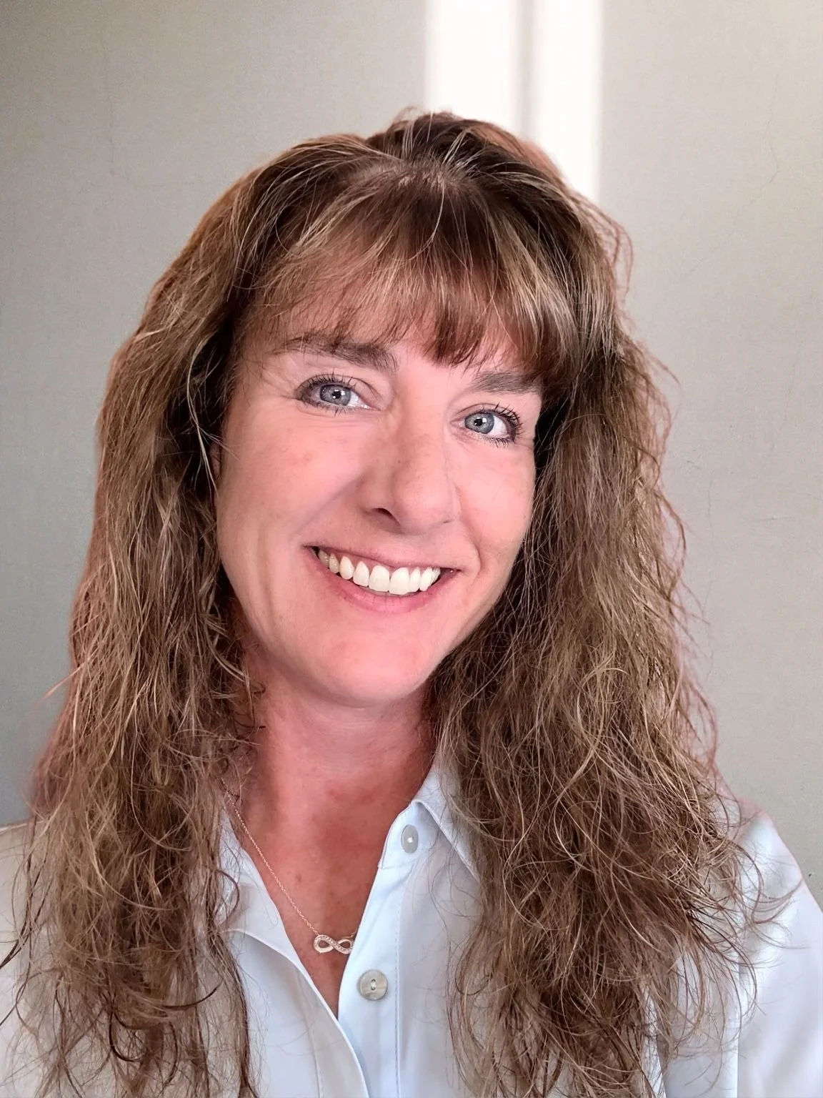A woman with curly, light brown hair and blue eyes, smiling, wearing a white collared shirt and a silver infinity necklace.