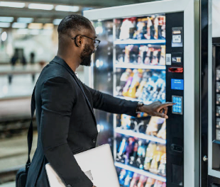 A man using a vending machine in an indoor setting, holding a laptop in one hand and pressing buttons on the vending machine with the other.