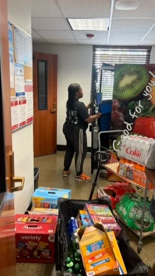 A woman shopping in a convenience store, with shelves of snacks and drinks, a cart, and promotional posters on the wall.