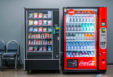 Vending machines side by side in a room with three empty chairs to the left.