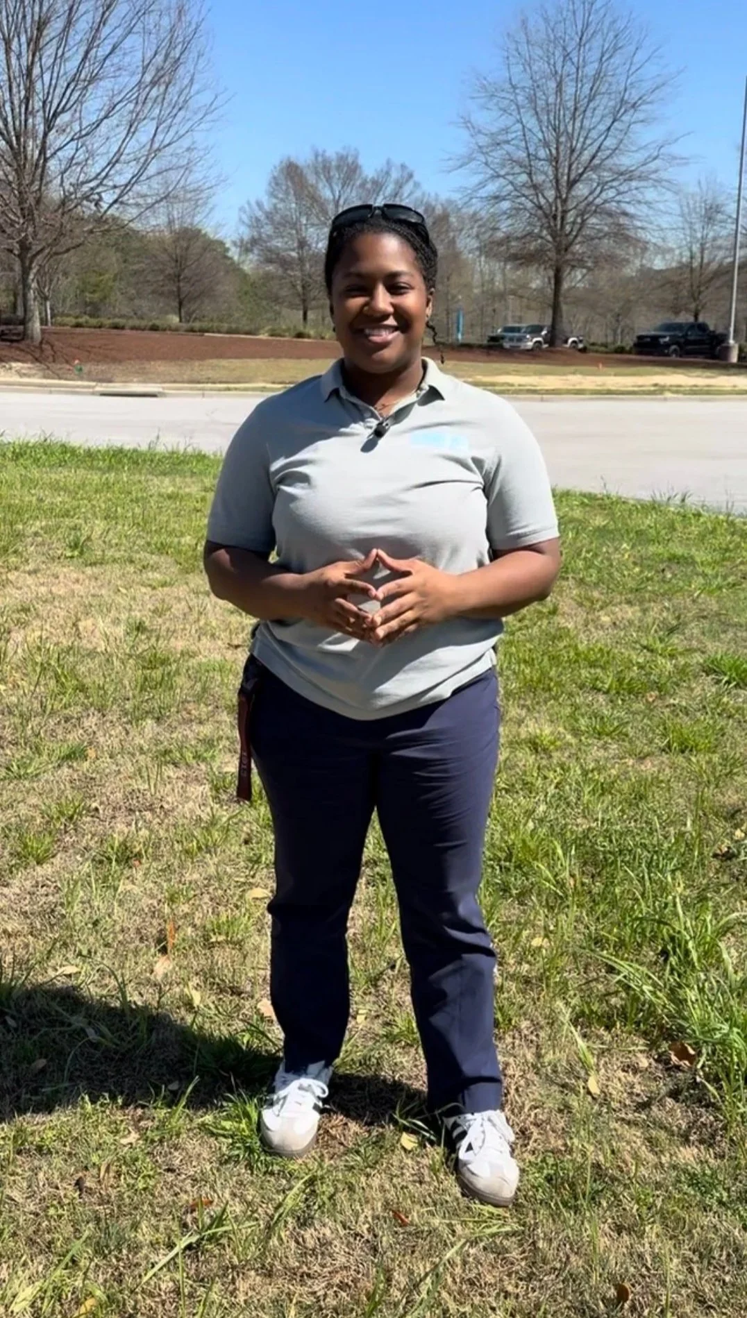 A woman smiling standing outdoors on a sunny day with leafless trees, a parking lot with several cars, and a grassy area in the background.