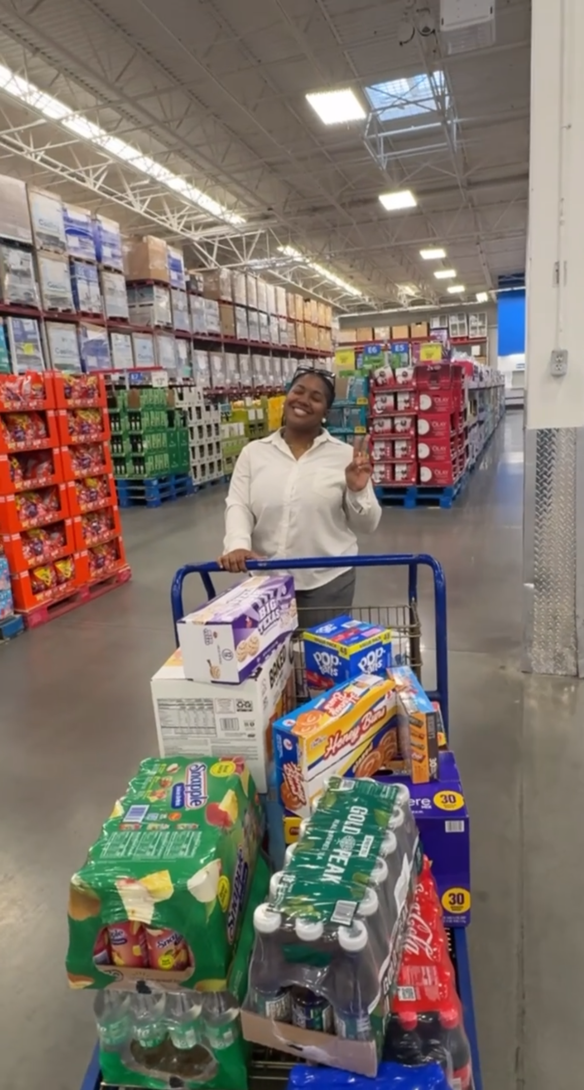 A woman smiling and making a peace sign while standing behind a shopping cart full of grocery items in a warehouse-style store.