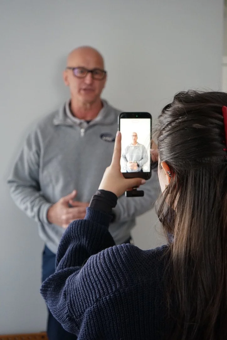A woman taking a photo of a man with her smartphone indoors. The man is wearing glasses and a gray jacket, standing against a plain wall.