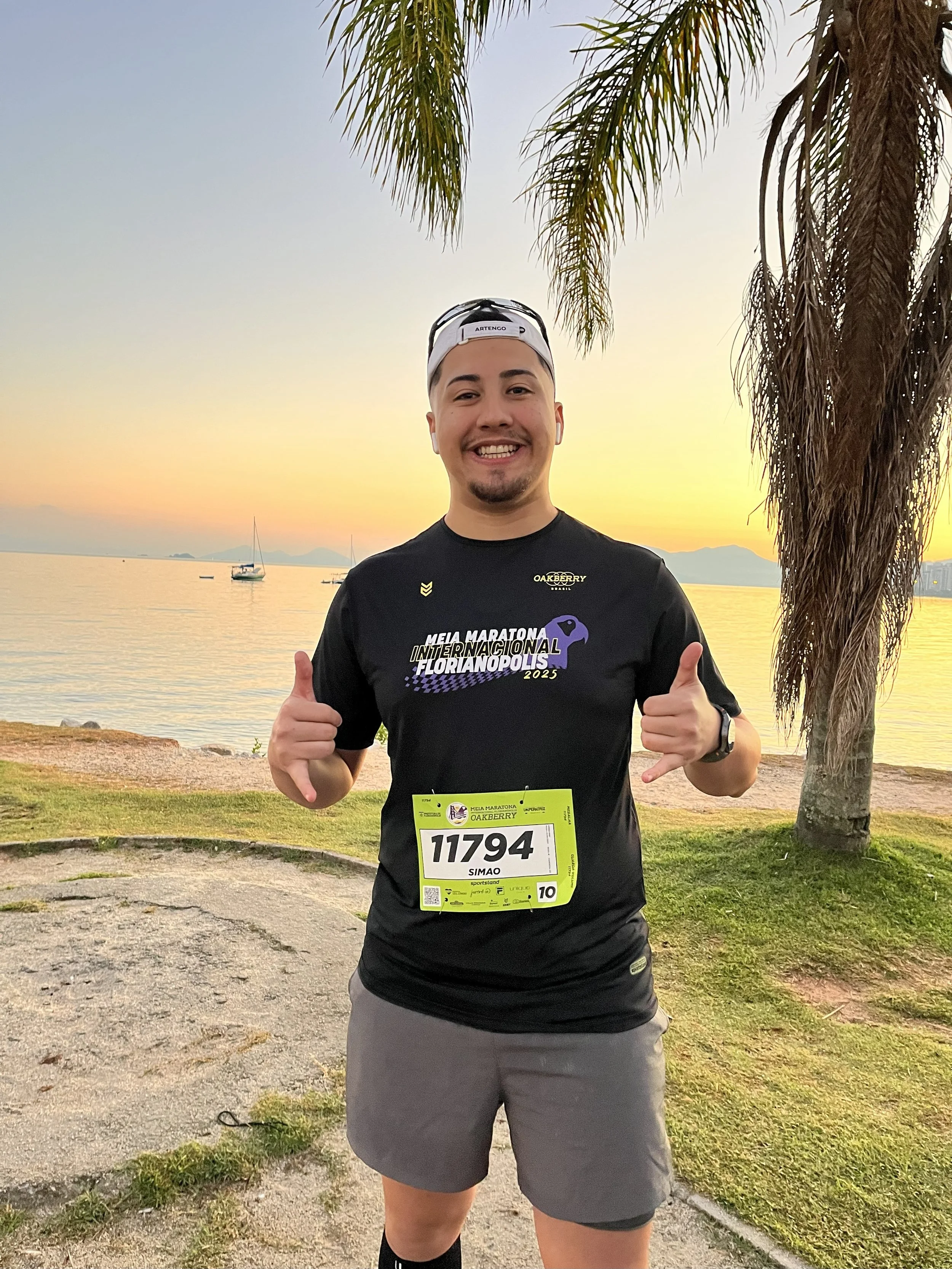 Homem sorridente vestindo roupa de corrida com número 11794, fazendo sinal de positivo com as mãos, ao pôr do sol na praia, com árvores ao fundo.