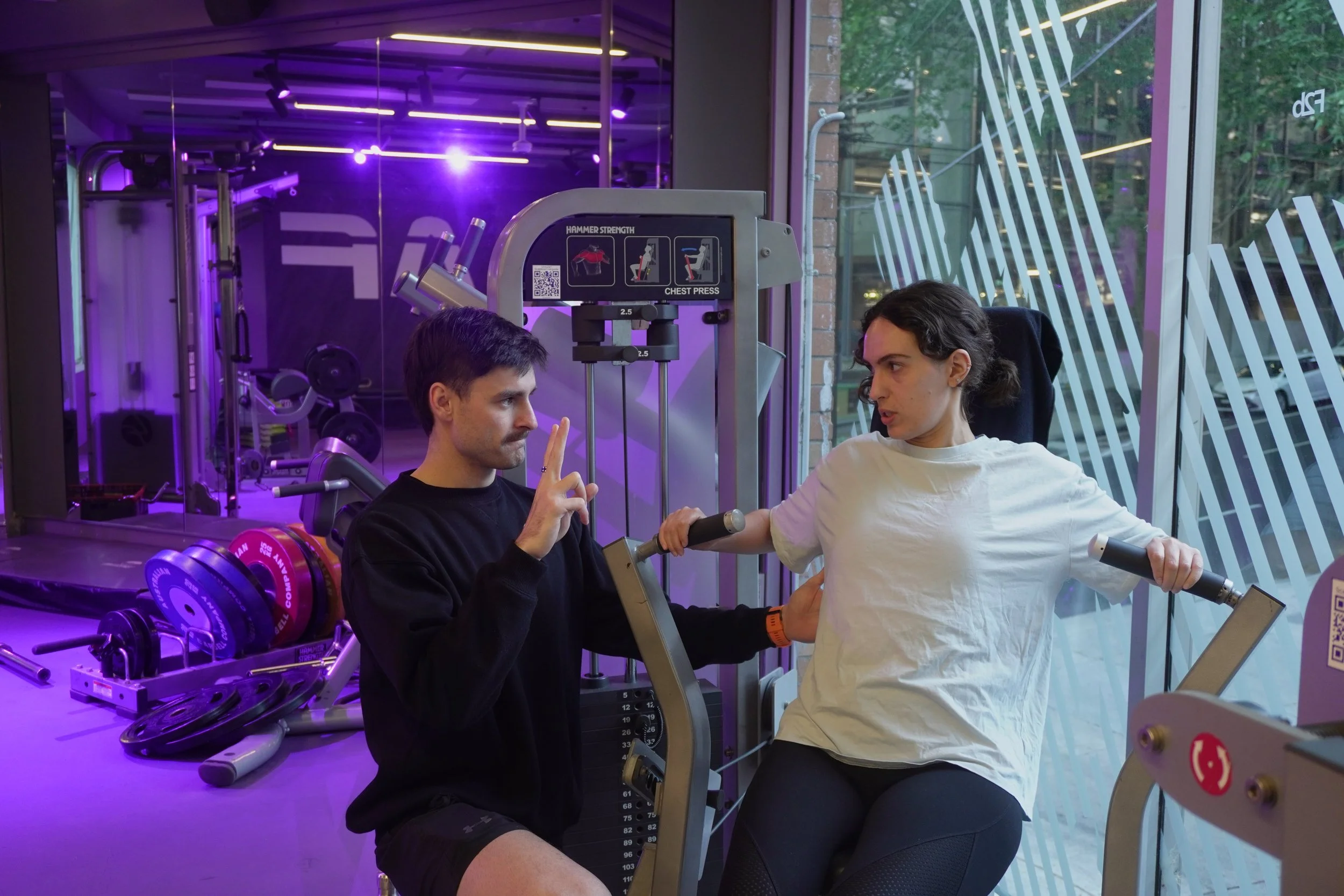 A personal trainer is instructing a woman using a seated chest press machine at a gym. The trainer is making a peace sign with his fingers while the woman is gripping the machine handles, with trees visible outside through large glass windows.