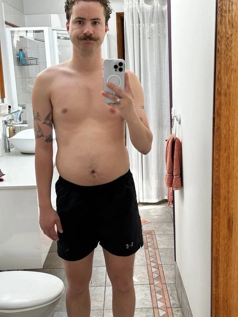 A shirtless man with a mustache taking a selfie in a bathroom mirror, wearing black Under Armour shorts. The bathroom has a shower with a glass door, a countertop with a sink, and a towel hanging on a rack. The man has tattoos on his left arm.