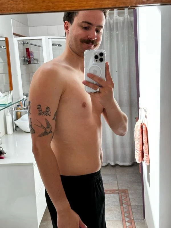 A shirtless man with a mustache taking a mirror selfie in a bathroom, wearing black shorts with tattoos on his left arm, standing in front of a mirror with a curtain and bathroom fixtures visible in the background.