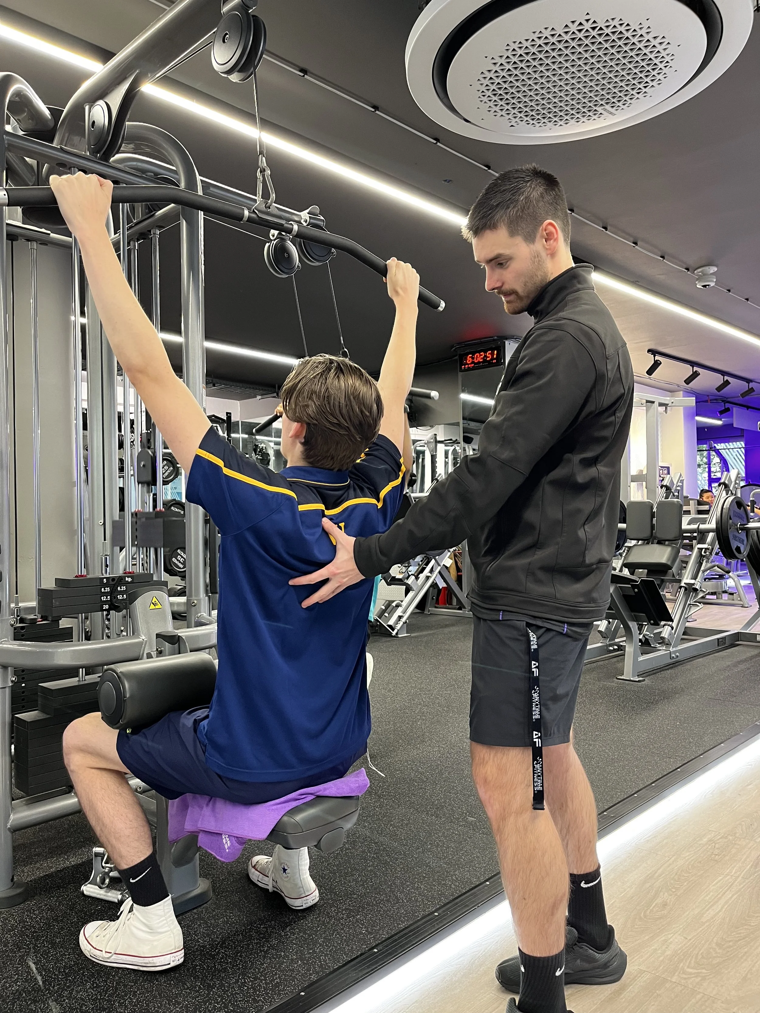 A young man with brown hair assisting a boy with light brown hair using gym equipment for an exercise in a modern gym, with other workout machines visible in the background.
