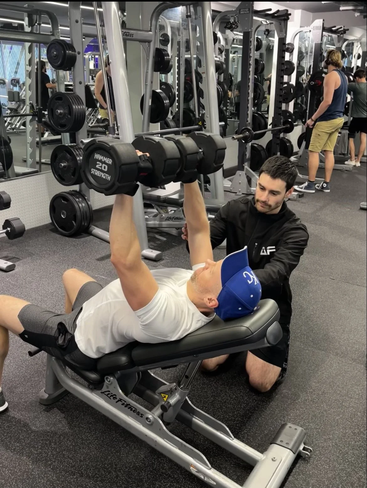 A man lying on a workout bench lifting dumbbells with a trainer assisting in a gym filled with exercise equipment.