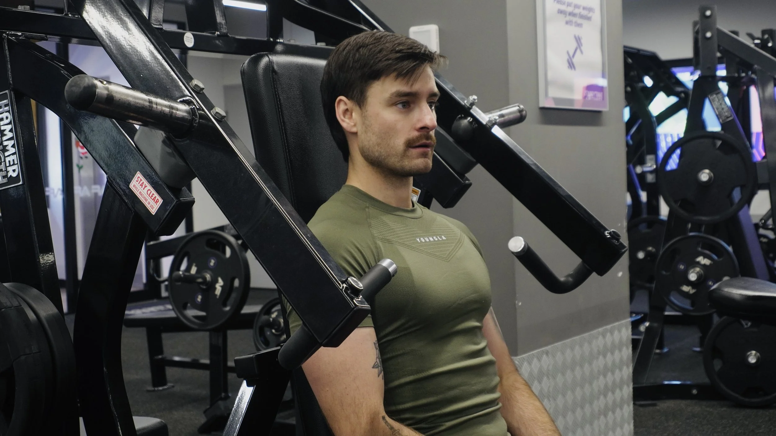 A man with dark hair and a mustache, wearing a green t-shirt, sitting on a seated chest press machine at the gym, with weight plates in the background.