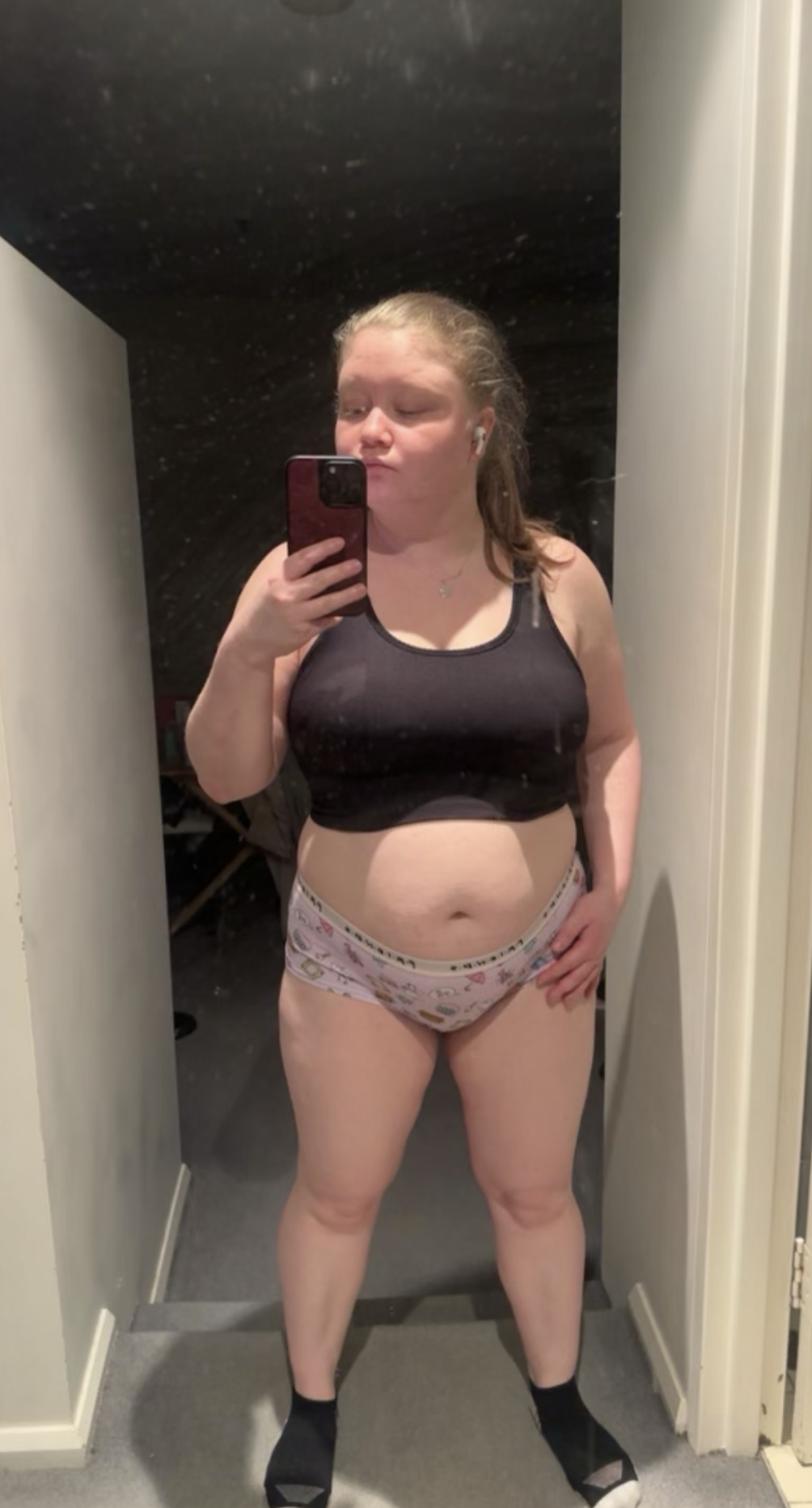 A woman taking a mirror selfie in a room, wearing a black sports bra and pajama bottoms with a pattern, with one hand on her hip and a neutral facial expression.