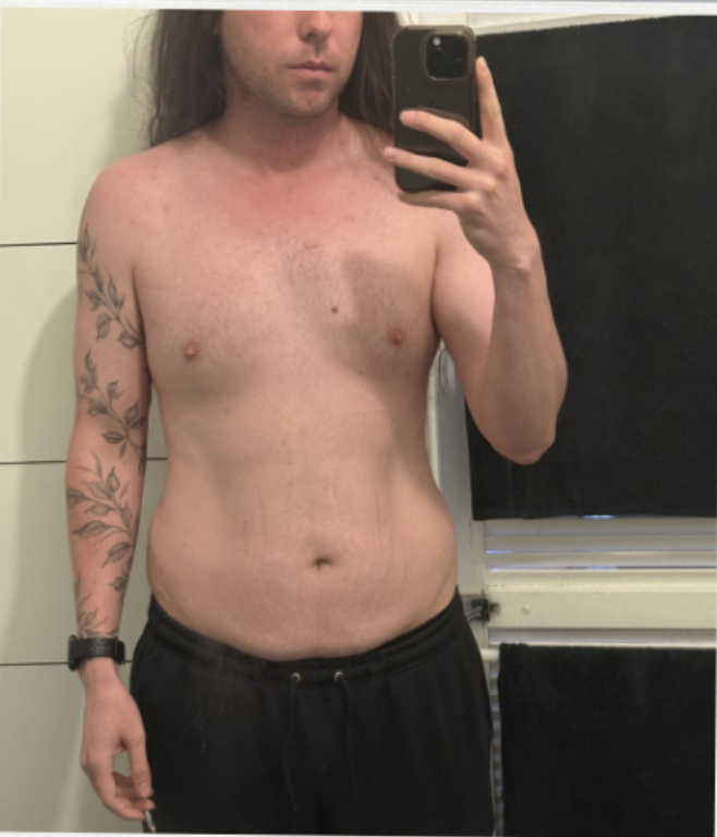 A shirtless person with long hair taking a mirror selfie in a bathroom, showing their torso and arm with a tattoo.