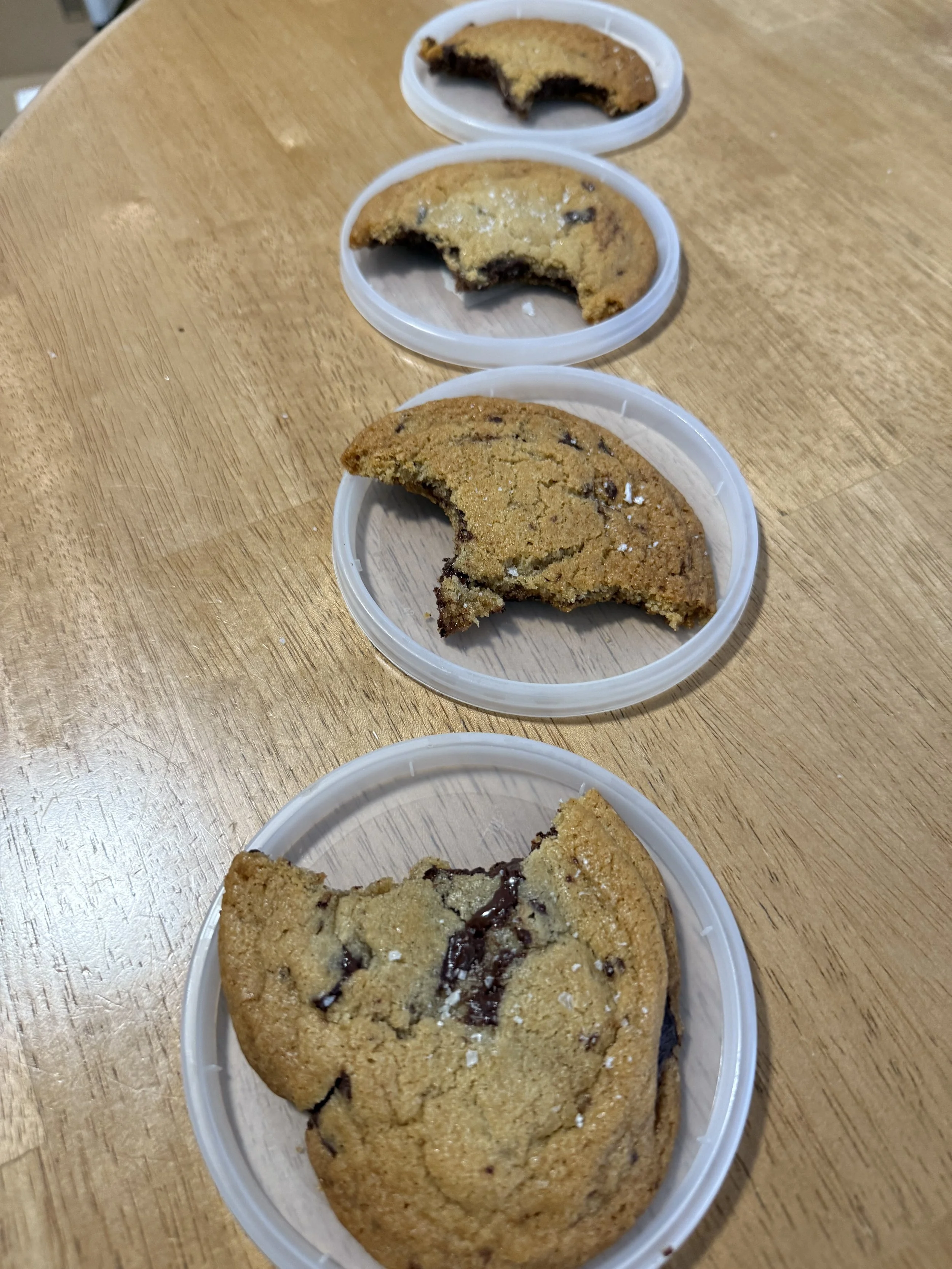 Four chocolate chip cookies in clear plastic containers arranged in a line on a wooden table, with the first cookie having a bite taken out of it.