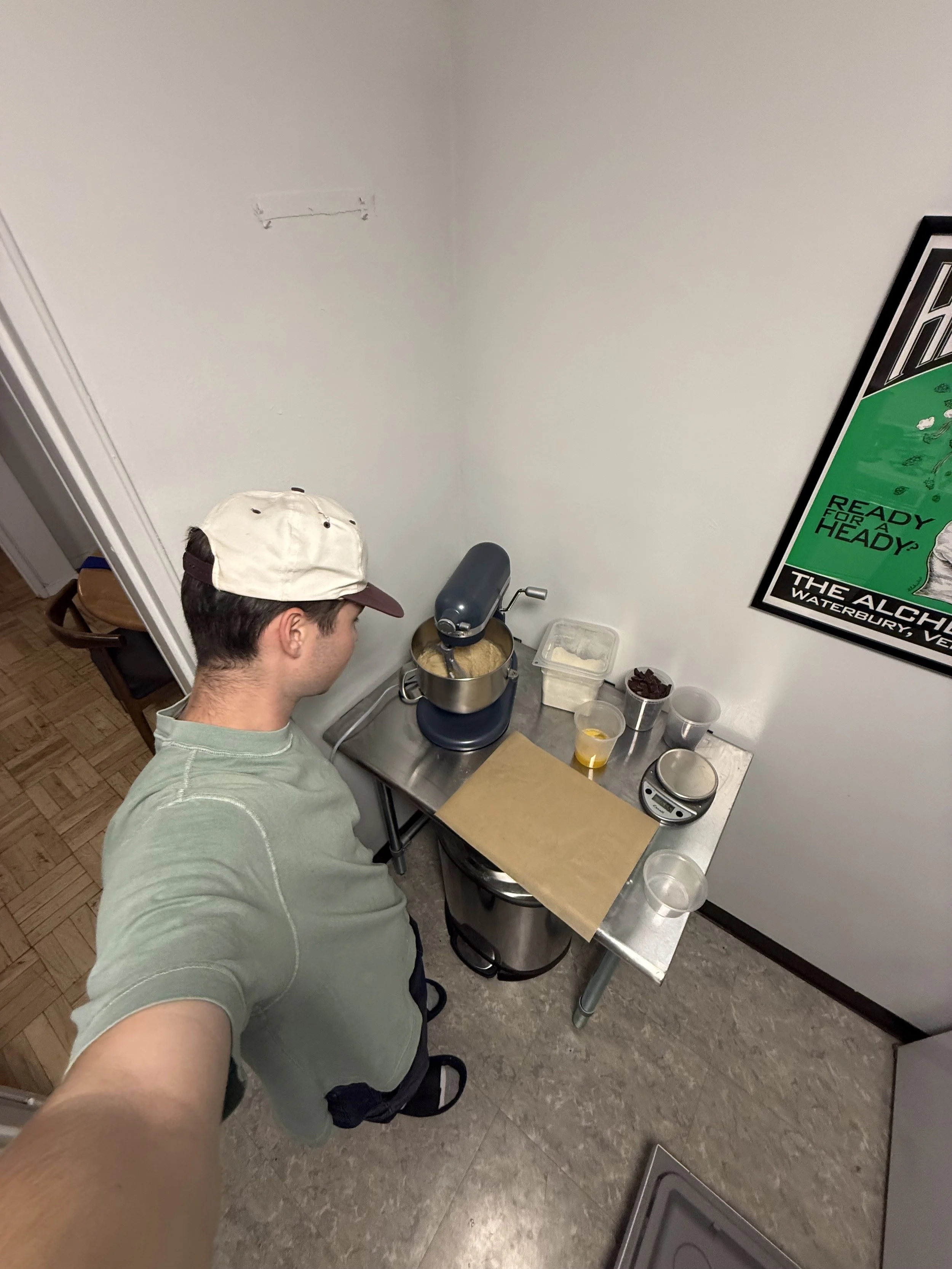 A person in a green shirt and beige cap is taking a selfie in a small kitchen or food prep area, where a stand mixer is mixing batter on a metal table, with various ingredients and containers around.