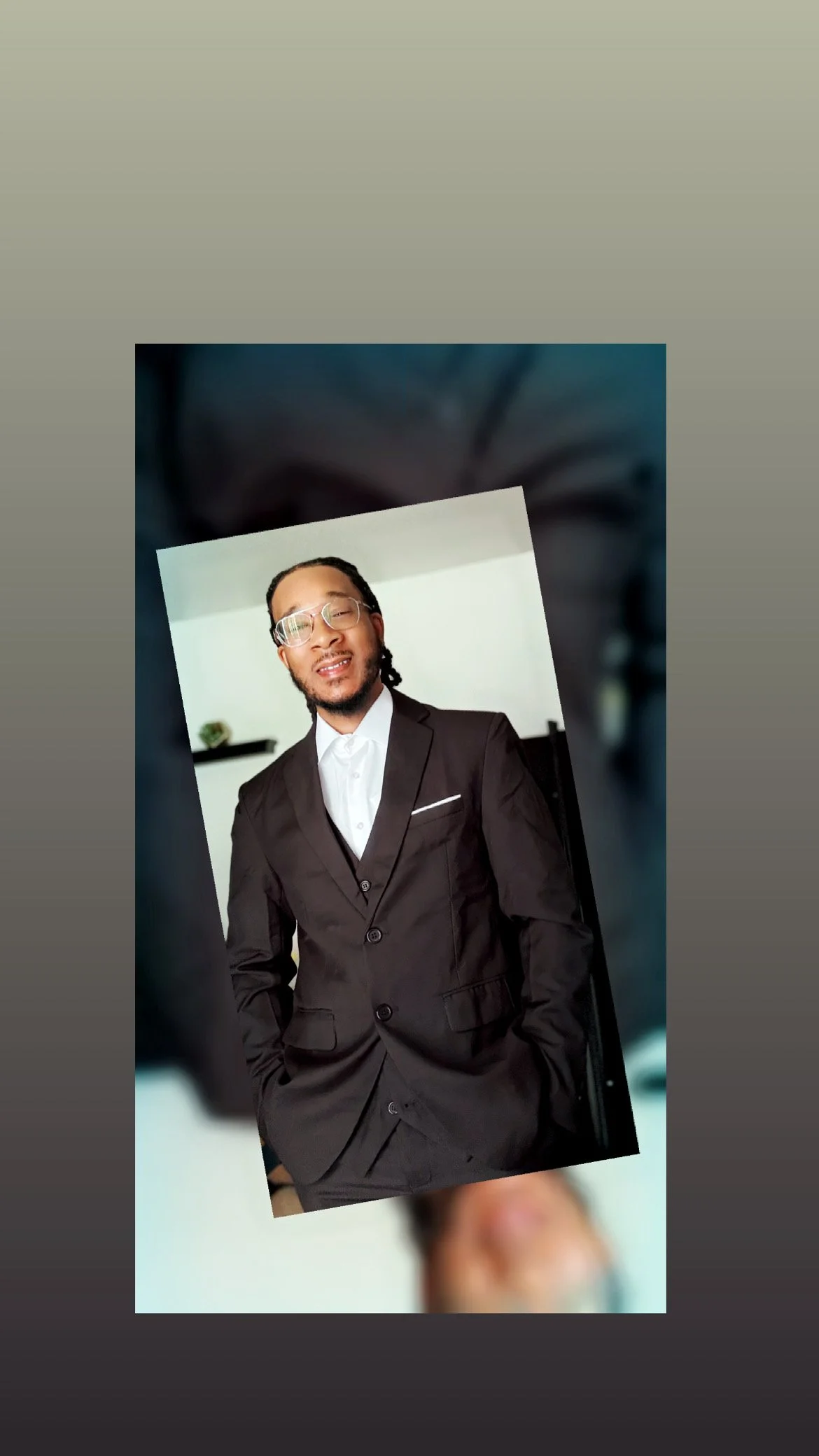 Portrait of a man wearing glasses and a black suit with a white shirt, standing indoors with a white background and shelves behind him.