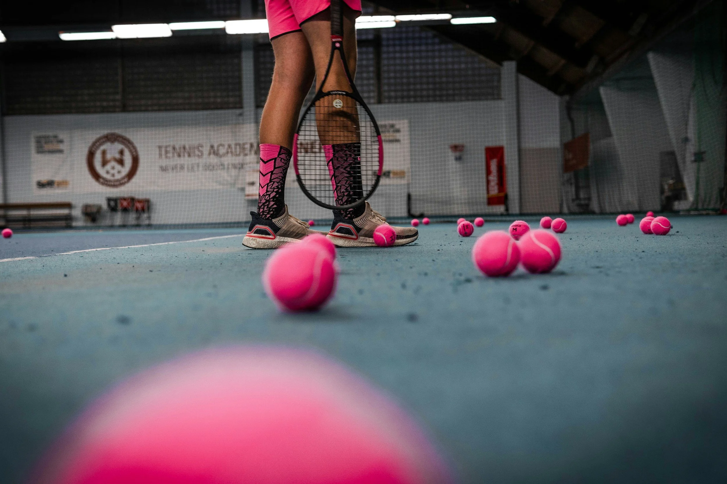 Person with pink and black patterned socks and beige tennis shoes holding a tennis racquet on an indoor tennis court, surrounded by pink tennis balls scattered on the court.