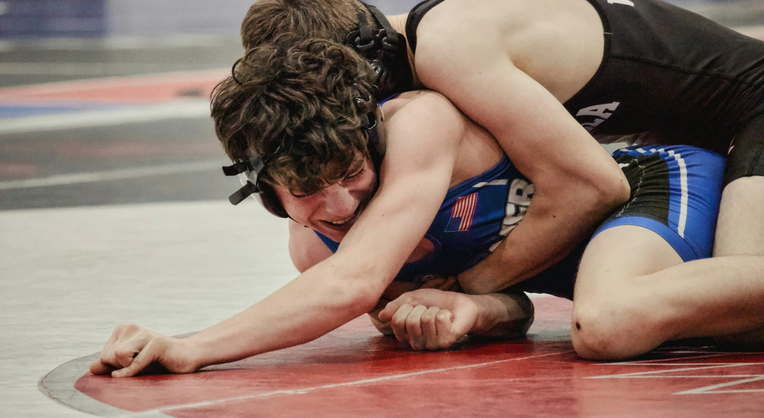 Two male wrestlers in a wrestling match, one pinned on the mat wearing a blue singlet with an American flag patch, and the other in a black singlet, on a red wrestling mat.