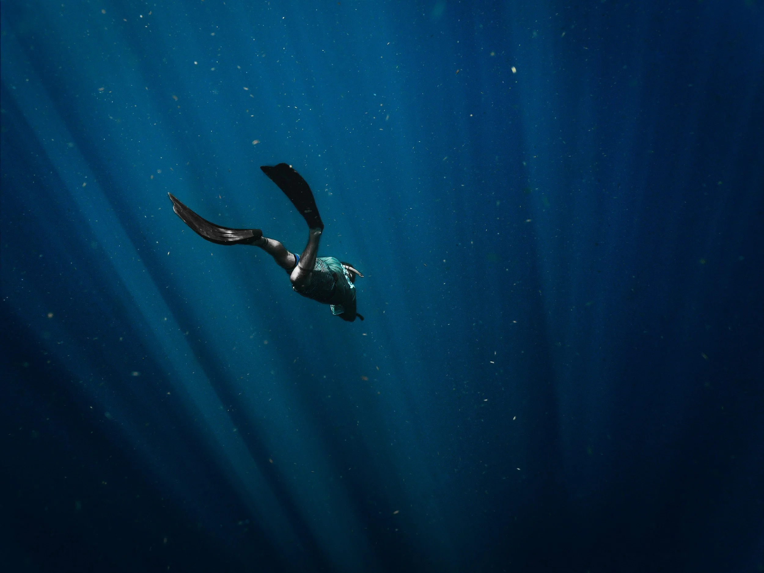 An underwater scene with a swimmer in a wetsuit swimming downward in the deep blue sea with sunlight rays penetrating the water.