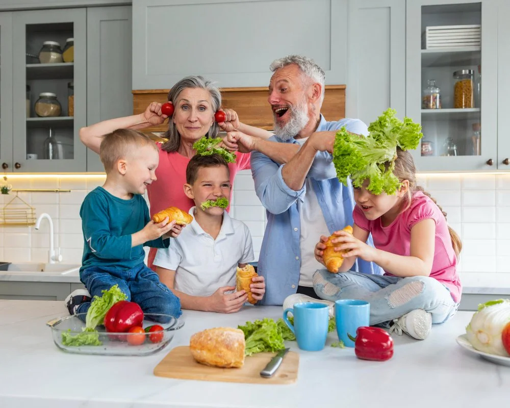 Family of six, including children and grandparents, playing and joking with vegetables and food in a modern kitchen.