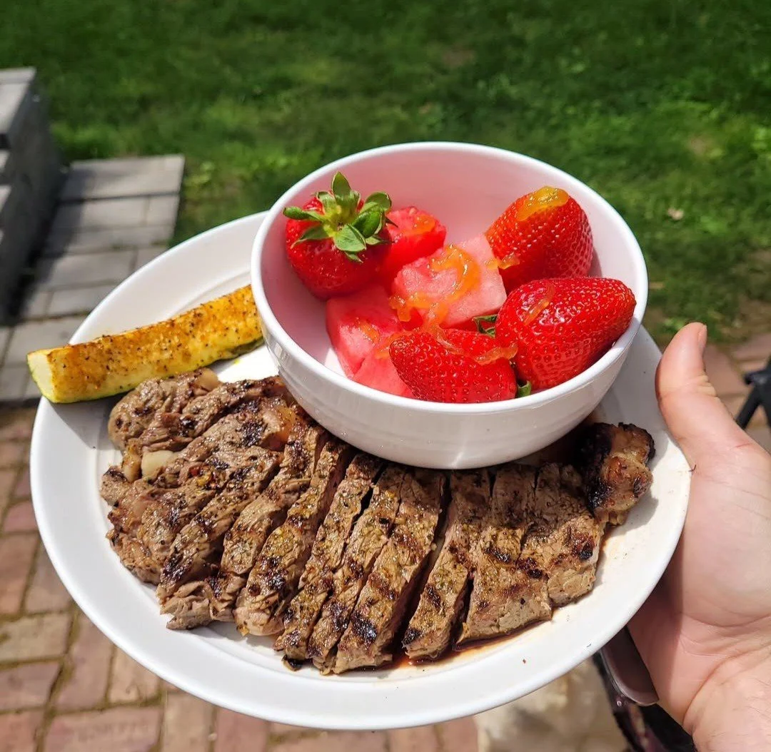 A white plate with grilled sliced steak, a grilled lemon wedge, and a bowl of fresh strawberries and sliced watermelon, held outdoors over a grassy area with a wooden deck in the background.