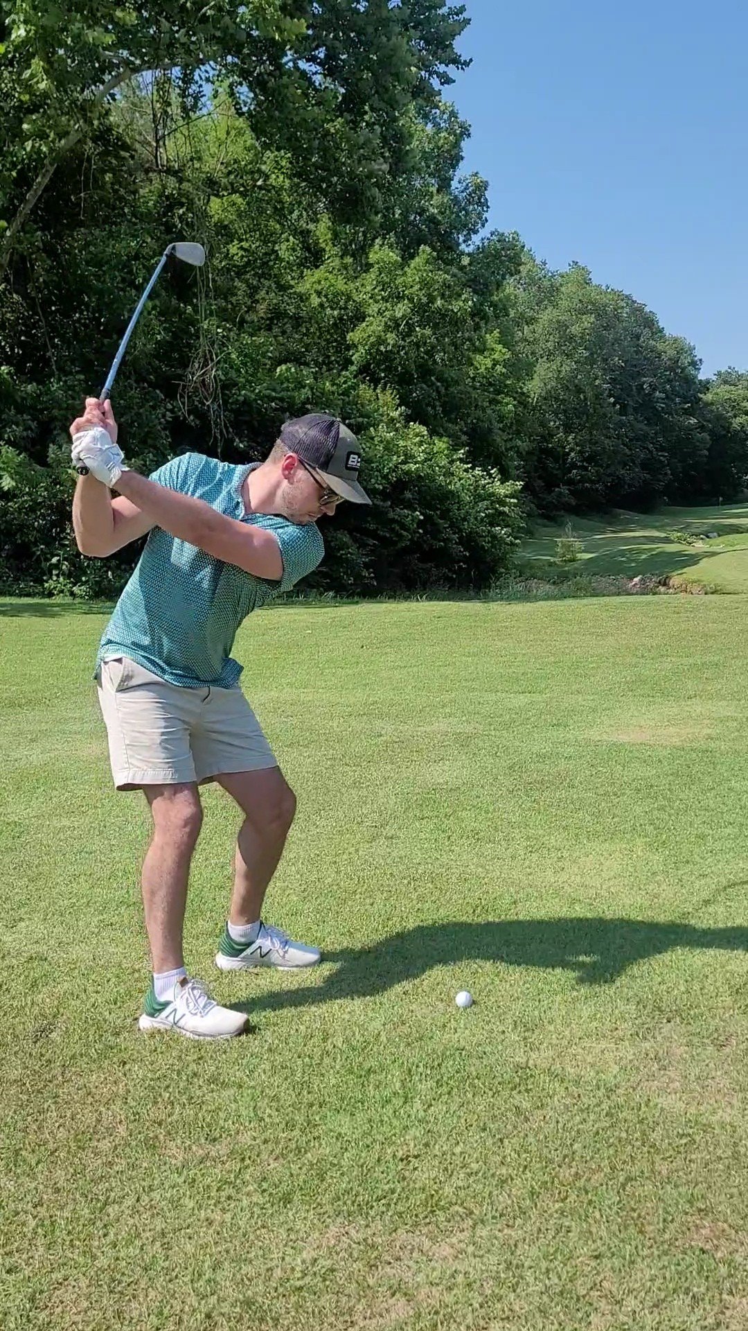A man wearing a cap, glasses, a blue shirt, beige shorts, and white shoes is preparing to hit a golf ball on a golf course. He is standing on the grass, holding a golf club, with a background of trees and blue sky.